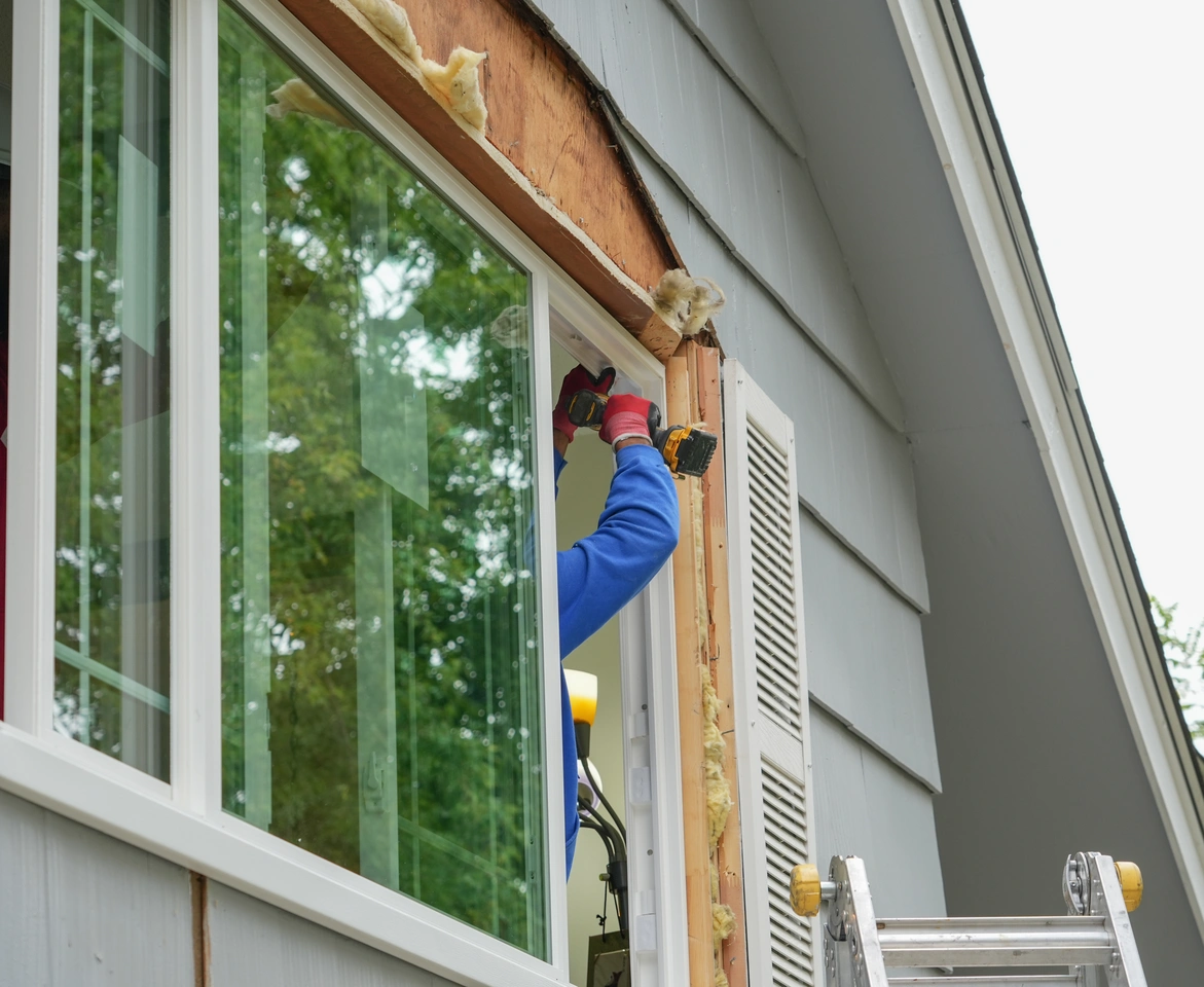 "Contractor installing energy efficient windows on a gray house exterior using a power drill, with insulation exposed and ladder positioned nearby.