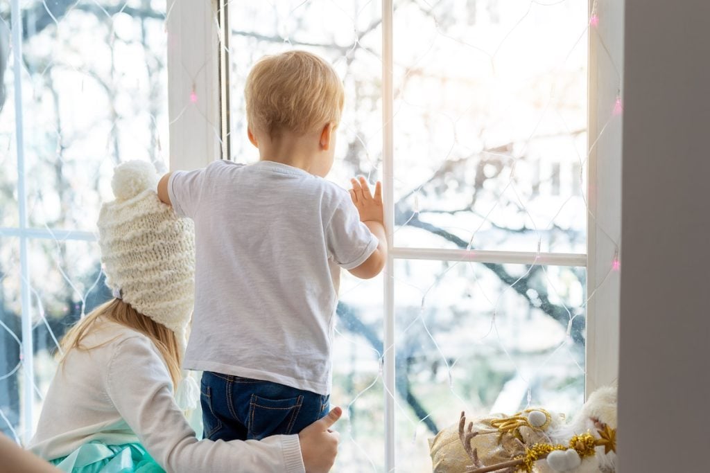 young brother and sister looking out of energy efficient windows decorated with holiday lights, staying warm indoors during winter.