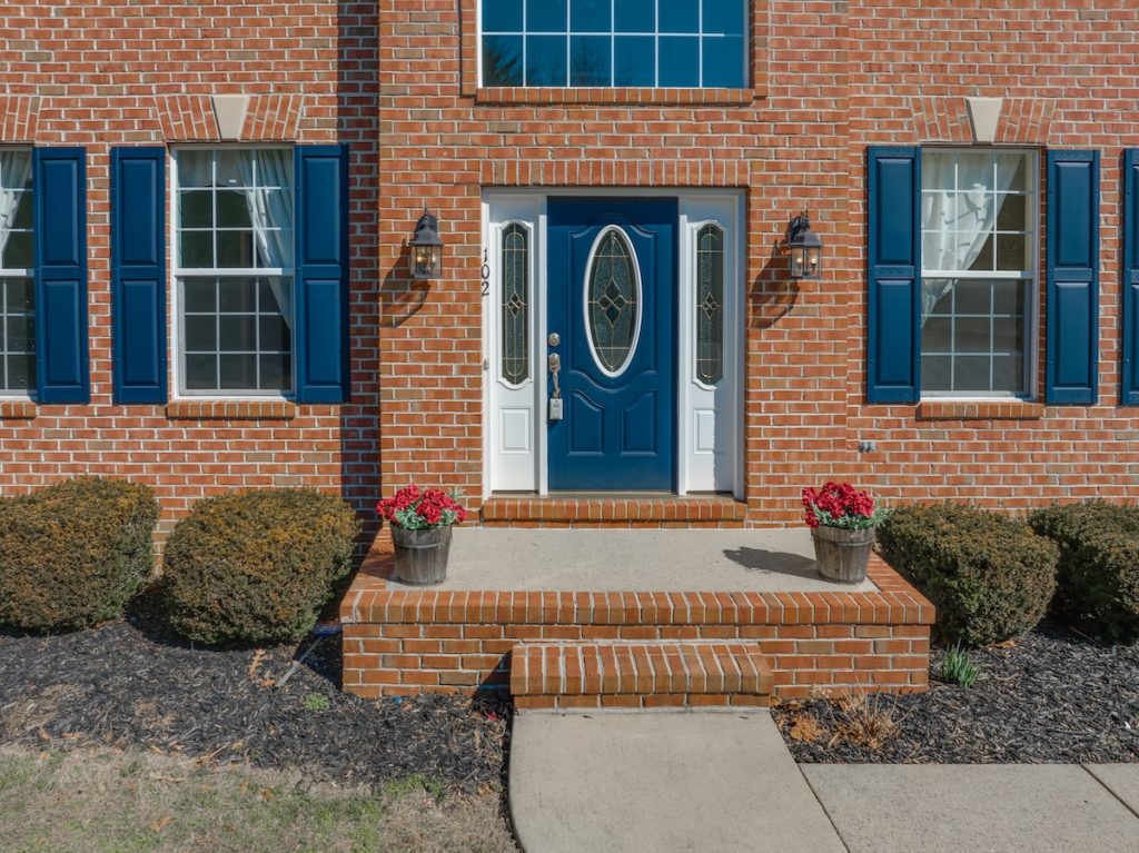 Close up view of elegant blue wooden door with oval stained window, with white sidelights, silver brass color door knob, iron wall light on red brick facade, red flower pot