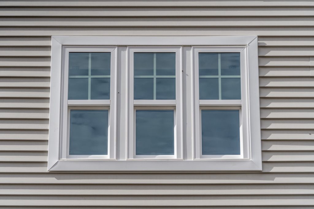 Triple hung window with fixed top sash and bottom sash that slides up, sash divided by white grilles a surrounded by white elegant frame  horizontal white vinyl siding on a new construction residence how to insulate windows for summer