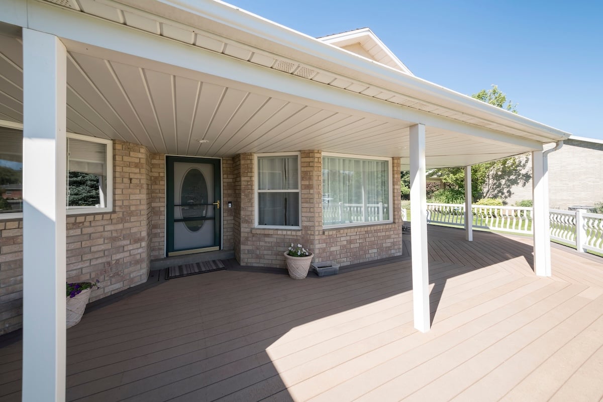 Wooden veranda of a house with screen door and white main door. Exterior of a house with bricks and two potted plants near the front door and a view of a lawn, house and trees on the right.