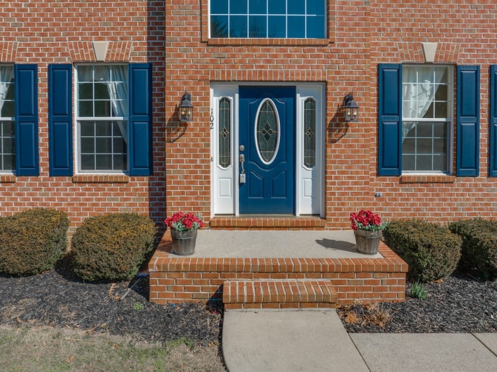 fiberglass vs steel door Close up view of elegant blue wooden door with oval stained window, with white sidelights, silver brass color door knob, iron wall light on red brick facade, red flower pot