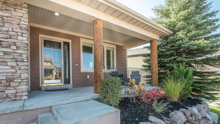 Pano Front exterior of a house with a raised edge landscape and stone brick walls. A porch with storm door, windows and log posts with a steel railings, and a concrete stairs near the garage with bricks. steel door