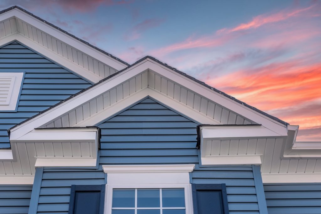 hardie board siding Triangle shape decorative gable with colonial white soffit and fascia on a blue horizontal vinyl siding modern American estate home with colorful dramatic stunning orange sunset sky