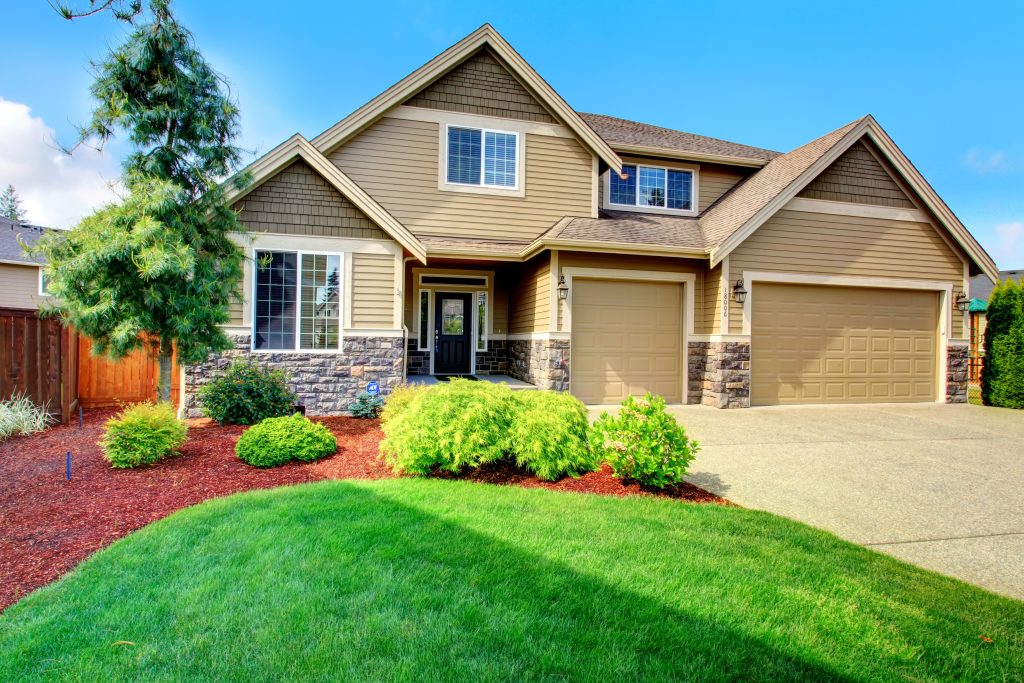 Clapboard siding house  with stone trim. View of  entrance porch, beautiful flower bed with green lawn, bushes and fir tree