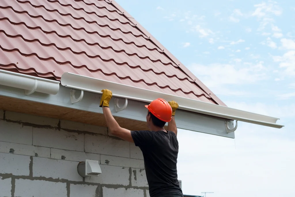 Worker in hard hat installing a gutter along the edge of a house roof with red metal shingles